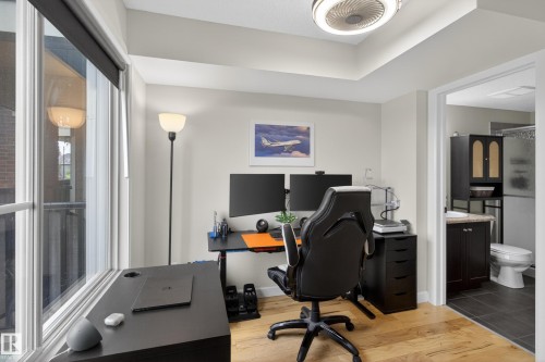 Spacious room featuring light wood-finish flooring, a tray ceiling with a modern flush-mount light fixture, and an expansive window with a roller shade - 211 3719 Whitelaw Lane, Edmonton, AB 