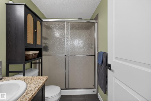 Bathroom featuring a shower with textured glass enclosure, a vanity with a light-colored speckled countertop, an integrated sink, and an over-the-toilet storage cabinet with textured door inserts - 211 3719 Whitelaw Lane, Edmonton, AB 
