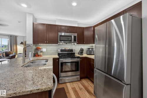 Kitchen featuring dark wood cabinetry, stainless steel appliances, light-toned granite countertops, and wood-finish flooring - 211 3719 Whitelaw Lane, Edmonton, AB 
