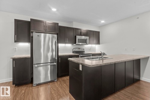 Kitchen featuring dark wood-finish cabinetry, stainless steel appliances, a center island with a double basin sink, light-toned countertops, and wood-finish flooring - 1302-1601 8 Avenue, Cold Lake, AB 