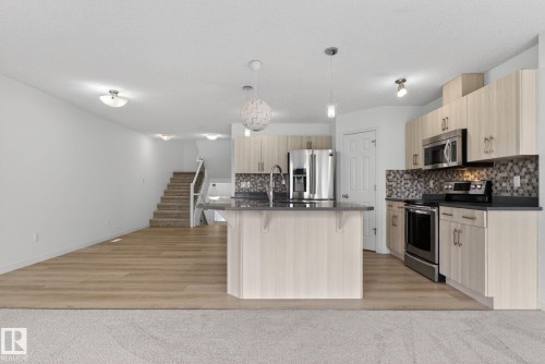Open-concept kitchen featuring light wood-finish cabinetry, stainless steel appliances, dark countertops, a decorative tile backsplash, and a central island with pendant lighting - 3806 Powell Wynd, Edmonton, AB 