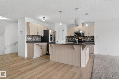 Open-concept kitchen featuring light-toned cabinetry, dark countertops, a mosaic tile backsplash, a central island with an undermount sink, and stainless steel appliances - 3806 Powell Wynd, Edmonton, AB 