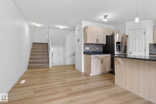 Open-concept living space featuring wood-finish flooring, a contemporary kitchen with light wood-finish cabinetry, dark countertops, and a mosaic tile backsplash - 3806 Powell Wynd, Edmonton, AB 