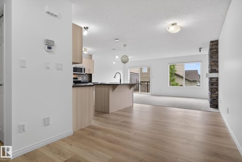Open-concept living space featuring wood-finish flooring extending into the kitchen, a contemporary kitchen island with a dark countertop, light wood cabinetry, a built-in microwave, and a stacked stone fireplace with a white mantel - 3806 Powell Wynd, Edmonton, AB 