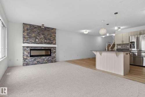 Open-concept living space featuring a stacked stone fireplace with a contemporary linear insert, light-toned wood-finish flooring, and a kitchen island with a double basin sink and pendant lighting - 3806 Powell Wynd, Edmonton, AB 
