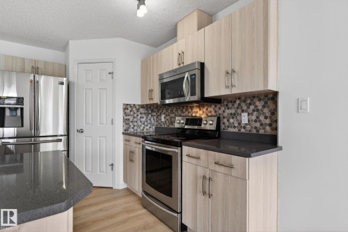 Kitchen featuring light wood-finish cabinetry, stainless steel appliances, dark countertops, a mosaic tile backsplash, and light wood-finish flooring - 3806 Powell Wynd, Edmonton, AB 