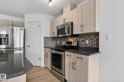 Kitchen featuring light wood-finish cabinetry, stainless steel appliances, dark countertops, a mosaic tile backsplash, and light wood-finish flooring - 