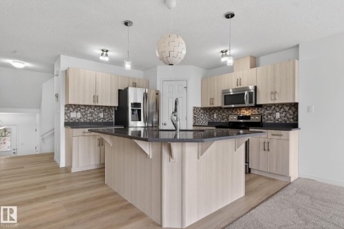 Kitchen featuring a central island with a dark countertop, light wood-finish cabinetry, a mosaic tile backsplash, and stainless steel appliances - 3806 Powell Wynd, Edmonton, AB 