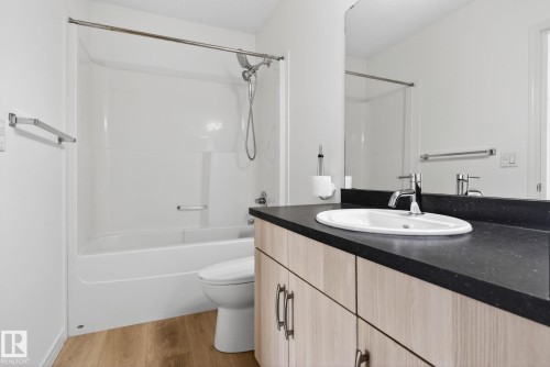 Bathroom featuring a white tub/shower combination with a chrome showerhead, a toilet, wood-finish flooring, a vanity with a dark countertop and an undermount sink, and a large wall-mounted mirror - 3806 Powell Wynd, Edmonton, AB 