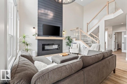 Open-concept living space featuring a two-story vaulted ceiling, a modern linear fireplace with a dark brick surround and shiplap accent wall, a glass-paneled staircase with natural wood handrails, large windows, and wood-finish flooring - 45 Elwyck Gate, Spruce Grove, AB 