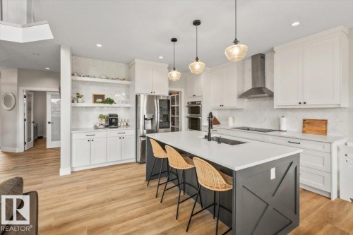 Contemporary kitchen featuring a large central island with seating, white shaker-style cabinetry, stainless steel appliances, wood-finish flooring, and pendant lighting - 45 Elwyck Gate, Spruce Grove, AB 