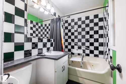 Bathroom featuring black and white checkerboard wall tile, a built-in bathtub, and a vanity with white cabinetry and a dark countertop - 5919 121 Avenue, Edmonton, AB 