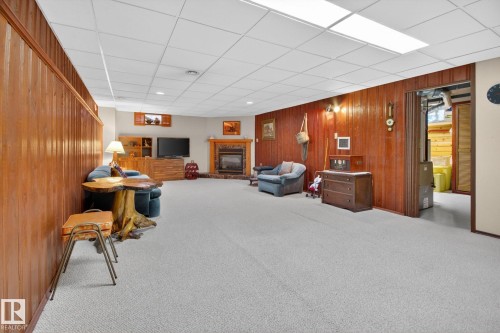 Spacious carpeted room featuring wood paneling, a stone fireplace with a wood mantle, and a drop ceiling with integrated lighting - 9243 74 Street, Edmonton, AB 