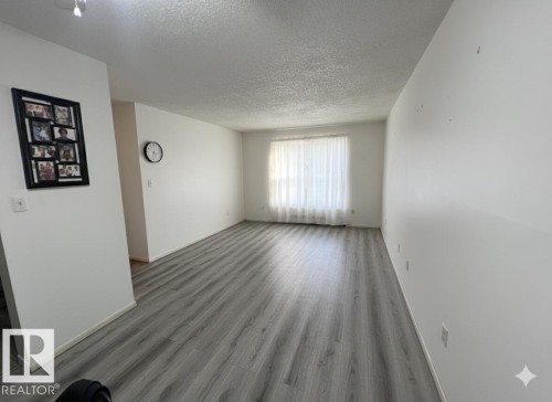 Spacious room featuring light gray wood-finish flooring, white walls, and a window with sheer white curtains - 212 2620 Mill Woods Road E, Edmonton, AB 