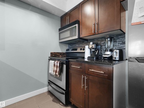 Kitchen featuring stainless steel appliances, rich wood-finish cabinetry, dark countertops, and a mosaic tile backsplash - 406 11820 22 Avenue, Edmonton, AB 