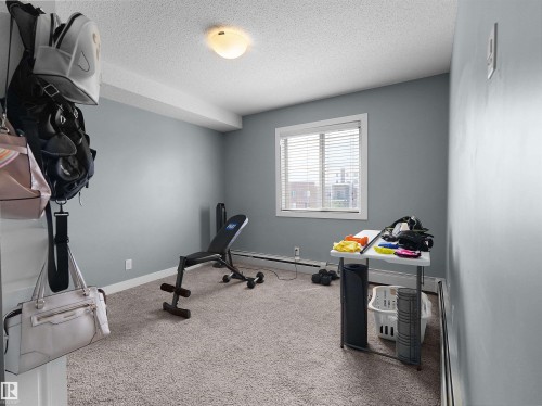 Light gray painted walls define this room featuring textured carpeting, a white trim window with blinds, and baseboard heating - 406 11820 22 Avenue, Edmonton, AB 