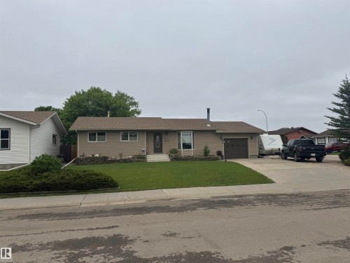 Ranch-style exterior featuring tan siding, a brown shingled roof, an attached garage, a paved driveway, and a front lawn with low-lying shrubbery - 4502 55 Avenue, Vegreville, AB 