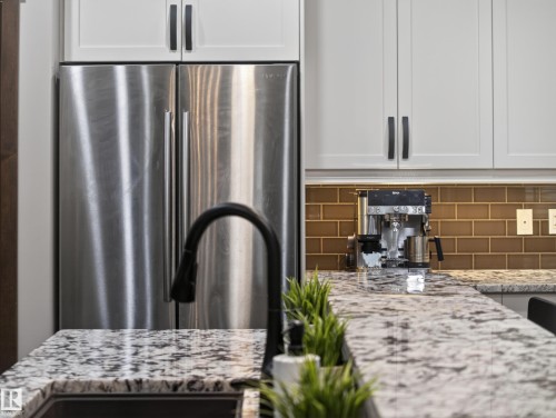 Stainless steel refrigerator integrated with white shaker-style cabinetry featuring matte black hardware, under-cabinet lighting, and a brown subway tile backsplash - 5608 Soleil Boulevard, Beaumont, AB 