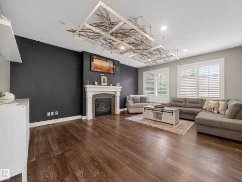 Spacious living area featuring rich wood-finish flooring, a contemporary fireplace with a decorative mantel, and two large windows fitted with plantation shutters - 5608 Soleil Boulevard, Beaumont, AB 
