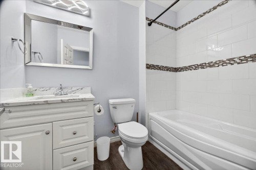 Bathroom featuring a white vanity with marble-look countertop, rectangular mirror, wood-finish flooring, and a tiled shower-tub combination with mosaic accent band - 1326 Cunningham Drive, Edmonton, AB 