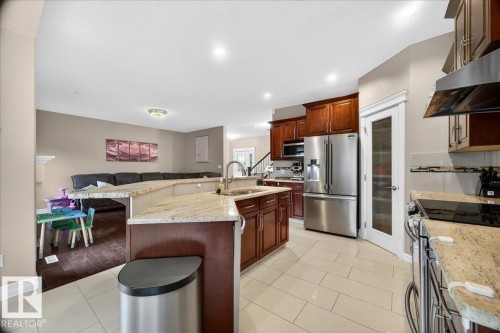Spacious kitchen featuring light tile flooring, granite countertops, cherry-toned cabinetry, stainless steel appliances, and an integrated kitchen island with a sink - 1326 Cunningham Drive, Edmonton, AB 
