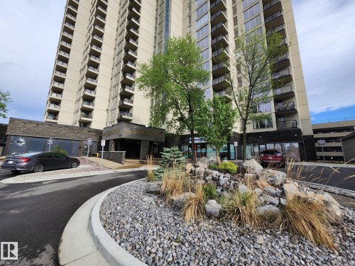 High-rise building with numerous balconies, featuring a stone-clad entrance, a rock garden with various plantings, and a paved circular driveway - 1600 10149 Saskatchewan Drive, Edmonton, AB 