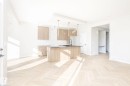 Open-concept kitchen and living area featuring wood-finish flooring, a kitchen island with a dark sink and matte black faucet, light wood cabinetry, a white tile backsplash, and multiple windows - 2206 Crossbill Link, Edmonton, AB 