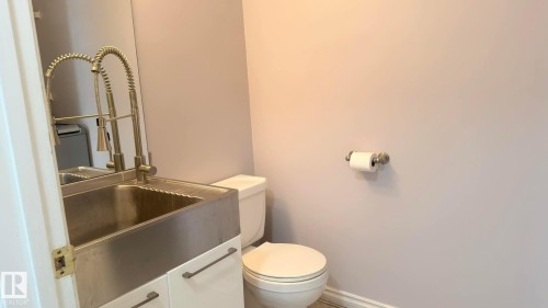 Powder room featuring a stainless steel apron front sink with a gooseneck pull-down faucet, white vanity cabinetry with linear hardware, and a white toilet. - 825 112B St, Edmonton, AB 