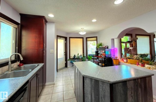 Kitchen featuring dark wood-finish cabinetry, a dual basin stainless steel sink with a gooseneck faucet, light-colored ceramic tile flooring, and a multi-level island with a wood-finish base and light-colored countertop overlooking an eat-in dining area. - 825 112B St, Edmonton, AB 
