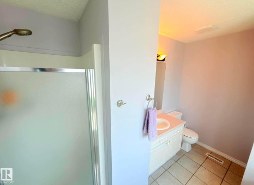 This Ensuite Bathroom featuring a shower with frosted glass door and a bronze-finish showerhead, a single vanity with a white cabinet and countertop, a white toilet, and tile flooring. - 825 112B St, Edmonton, AB 