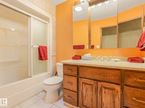 Bathroom featuring a wood-finish vanity with an integrated sink and light-toned countertop, a three-door mirrored medicine cabinet, a full-size toilet, and a combination tub and shower with sliding glass doors - 11921 231 Street, Edmonton, AB 