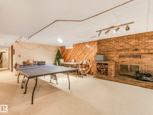 Spacious recreational room featuring a brick fireplace with a wood storage cubby, track lighting, light-toned carpet flooring, and a wood-paneled accent wall - 11921 231 Street, Edmonton, AB 