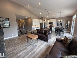 Open-concept living space featuring wood-finish flooring, a vaulted ceiling, recessed lighting, and a contemporary kitchen with white cabinetry and dark countertops - 