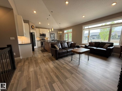 Open-concept living space featuring wood-finish flooring, recessed lighting, expansive windows, a kitchen island with pendant lighting, and white cabinetry with stainless steel appliances - 2 104 Allard Link, Edmonton, AB 