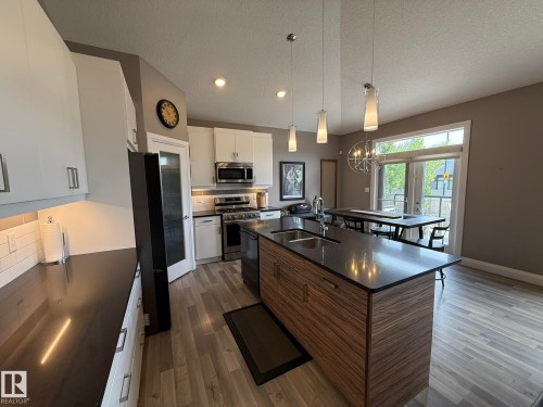 Modern kitchen featuring an island with wood-finish cabinetry and a dark countertop, stainless steel appliances, white subway tile backsplash, and light wood-finish flooring - 2 104 Allard Link, Edmonton, AB 