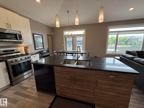 Open-concept kitchen featuring an island with a double basin sink, dark countertop, and wood-finish cabinetry - 2 104 Allard Link, Edmonton, AB 