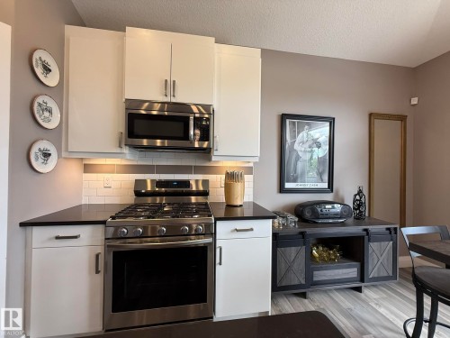 Kitchen area featuring white cabinetry, dark countertops, a stainless steel gas range, a coordinating microwave, and a subway tile backsplash - 2 104 Allard Link, Edmonton, AB 