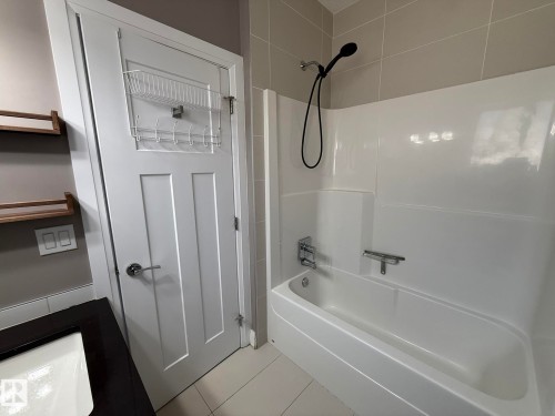 Bathroom featuring a white bathtub and shower combination with a black shower head, light grey wall tiles, and white floor tiles - 2 104 Allard Link, Edmonton, AB 