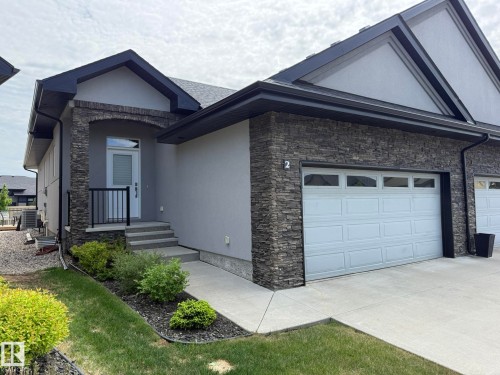 Modern exterior featuring a stone facade, a single garage door with upper window panels, a front entry with a black railing, a concrete walkway, and landscaped garden beds - 2 104 Allard Link, Edmonton, AB 