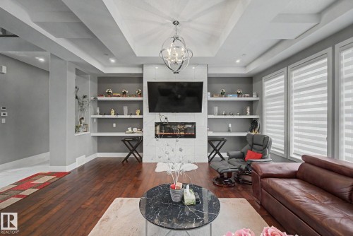 Spacious living area featuring engineered wood flooring, a tiered ceiling with recessed lighting and a central chandelier, and a contemporary fireplace with a television niche - 3036 59 Avenue, Rural Leduc County, AB 