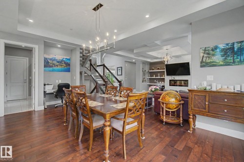 Open-concept living area featuring rich wood-finish flooring, a contemporary linear chandelier, tray ceiling details, a built-in electric fireplace, and a striking black open-riser staircase - 3036 59 Avenue, Rural Leduc County, AB 