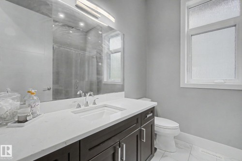 Bathroom featuring a dark wood-finish vanity with a white countertop and undermount sink, a large mirror with integrated lighting, a white toilet, and a window with frosted glass - 3036 59 Avenue, Rural Leduc County, AB 