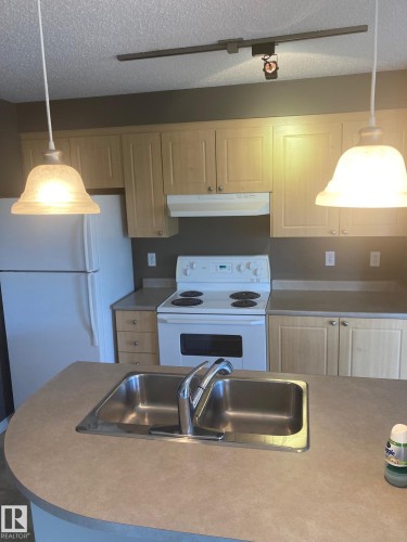 Kitchen featuring a double basin stainless steel sink, light wood-finish cabinetry, white electric range, white refrigerator, and track lighting - #206 616 Mcallister Lo Sw, Edmonton, AB - Indoor Photo Showing Kitchen With Double Sink