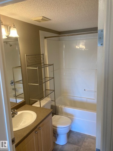 Bathroom featuring a light wood-finish vanity with an integrated sink, a neutral countertop, and a white bathtub with shower - #206 616 Mcallister Lo Sw, Edmonton, AB - Indoor Photo Showing Bathroom
