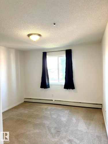 Carpeted room featuring a double window with dark curtains, baseboard heating, a ceiling-mounted light fixture, and textured ceiling finish - 312 2305 35A Avenue, Edmonton, AB 