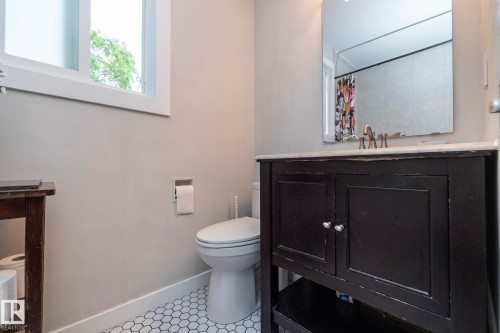 Bathroom featuring a dark wood vanity with a light-toned countertop, a rectangular mirror, a white toilet, and white hexagonal floor tiling - 7719 86 Avenue, Edmonton, AB 