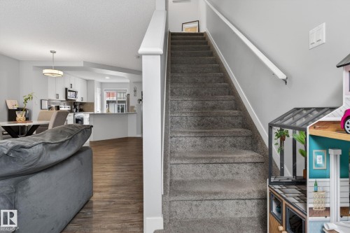 Carpeted staircase with white handrail and baseboards, opening to a living area with wood-finish flooring and a kitchen featuring white cabinetry, stainless steel appliances, and a peninsula with countertop seating - 62 3305 Orchards Link, Edmonton, AB 