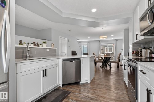 Modern kitchen featuring white shaker cabinetry, stainless steel appliances, a grey subway tile backsplash, and wood-finish flooring - 62 3305 Orchards Link, Edmonton, AB 