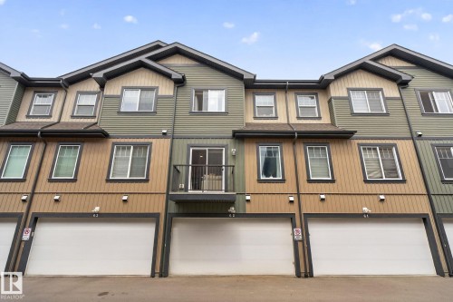 Modern multi-level townhome featuring a mix of horizontal siding and vertical paneling, an integrated single-car garage, a second-story balcony with metal railing, and multiple windows with dark trim - 62 3305 Orchards Link, Edmonton, AB 