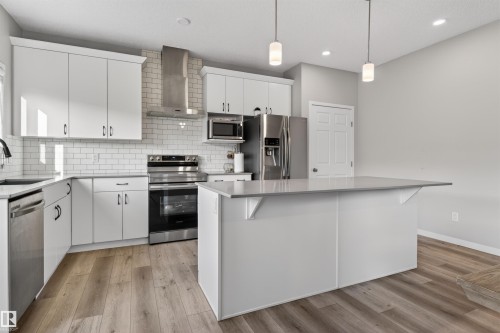 Modern kitchen featuring an expansive island with a light-toned countertop, white cabinetry with dark hardware, stainless steel appliances, a subway tile backsplash, and wood-finish flooring - 22638 99A Avenue, Edmonton, AB 
